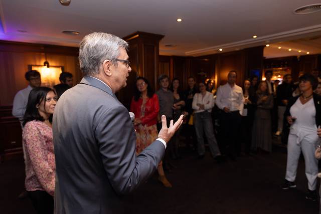 A man in a suit speaks in front of a group of attentive people at an indoor event.