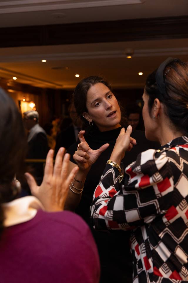 A woman is gesturing while talking to two other people at an indoor gathering.