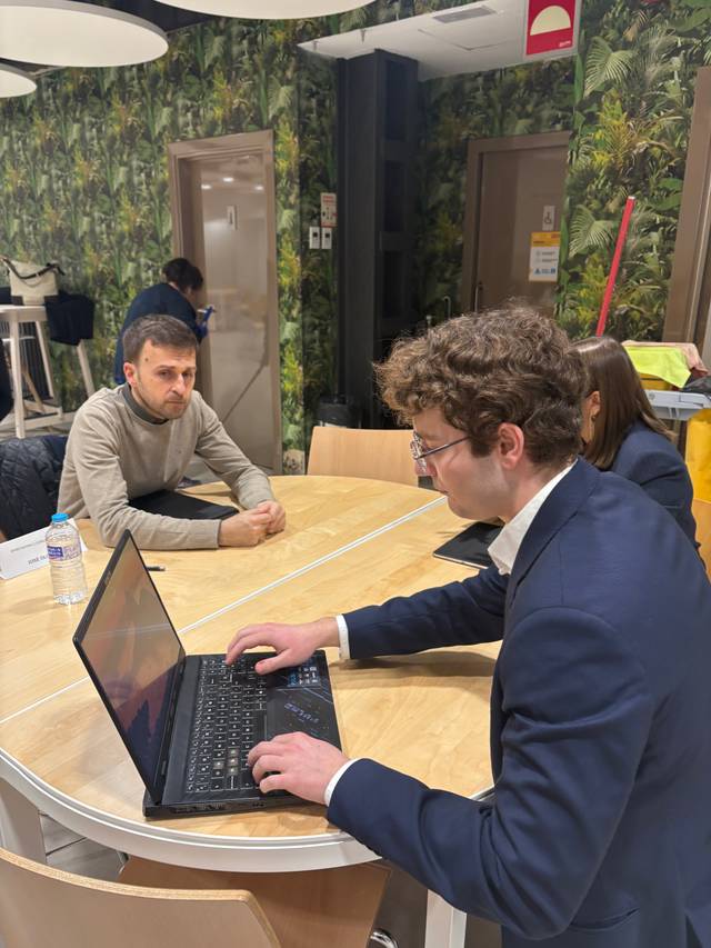 Dos personas están sentadas en una mesa, una de ellas trabajando en un portátil mientras la otra observa.