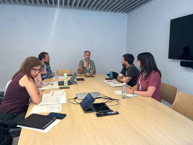 Un grupo de seis personas participa en una reunión en una sala con mesas de trabajo.