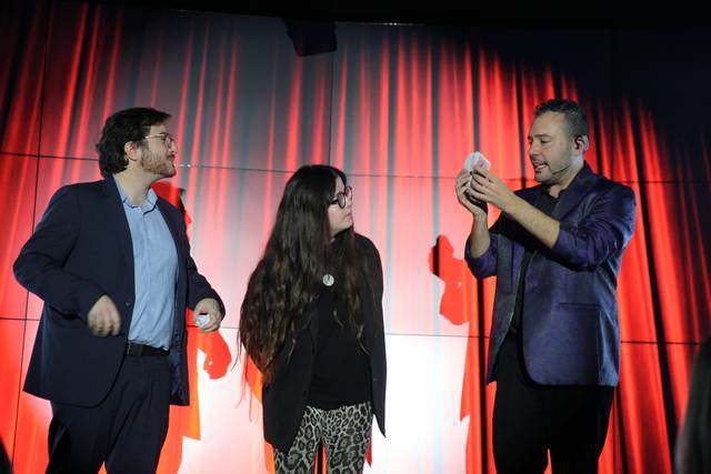 Tres personas sobre un escenario, dos hombres y una mujer, observan un acto durante un espectáculo de magia.