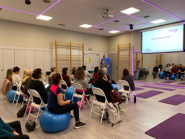 Un grupo de personas asiste a una charla en un salón con equipo de ejercicio y pantallas.