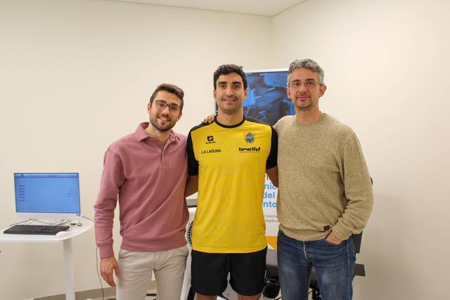 Tres hombres sonriendo a la cámara, uno de ellos vestido con una camiseta deportiva amarilla.