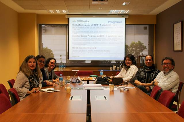 Un grupo de seis personas sentadas alrededor de una mesa de conferencia en una sala con una pantalla proyectando una presentación titulada 'Programs'.