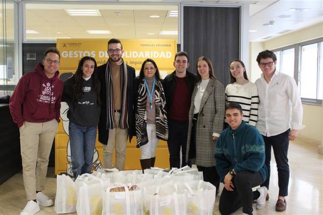 Un grupo de personas posa sonriente con bolsas de alimentos en un entorno interior frente a un cartel de 'Gestos Solidaridad'.