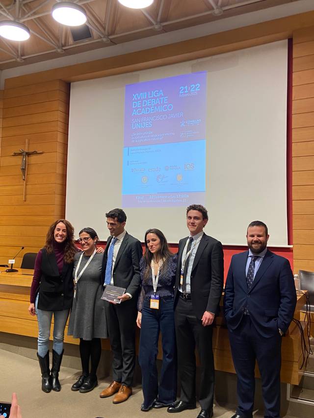 Un grupo de seis personas posando en un evento académico frente a un cartel que anuncia la XVIII Liga de Debate Académico UNIRE.
