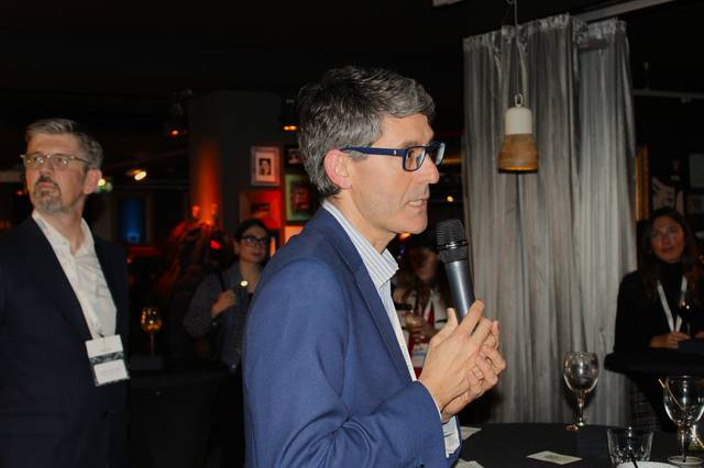 Un hombre con gafas y chaqueta azul está hablando en un micrófono durante un evento en un lugar con iluminación tenue.