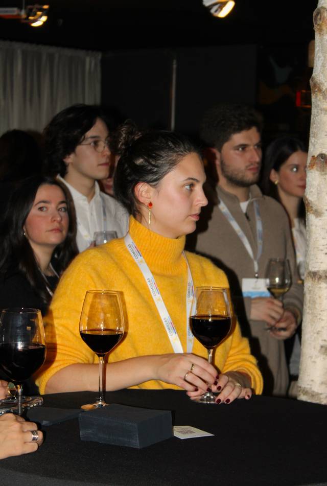 Mujer con suéter amarillo sostiene una copa de vino en un evento.