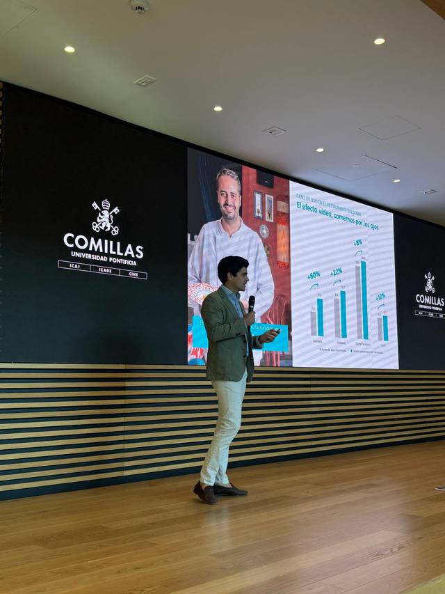 Un presentador habla frente a una gran pantalla con gráficas en un evento en la Universidad de Comillas.