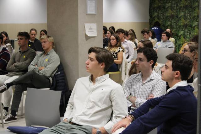 A group of young adults sitting in a seminar room, attentively listening to a speaker out of view.