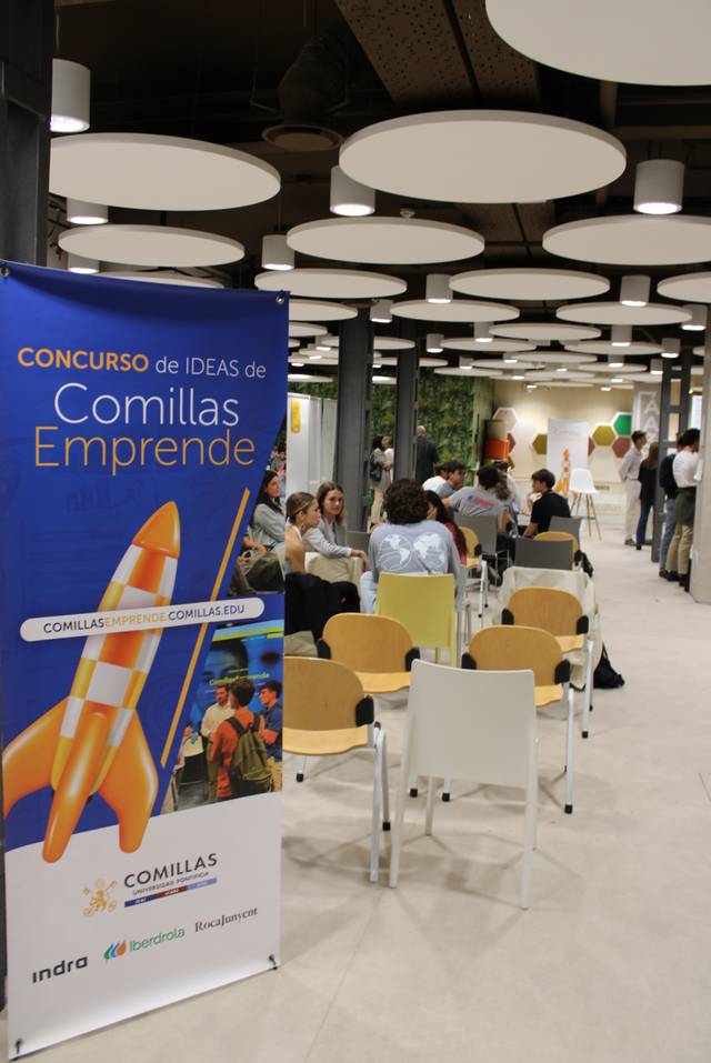 An indoor event at Comillas Emprende featuring a banner, chairs, and people engaging in conversations.