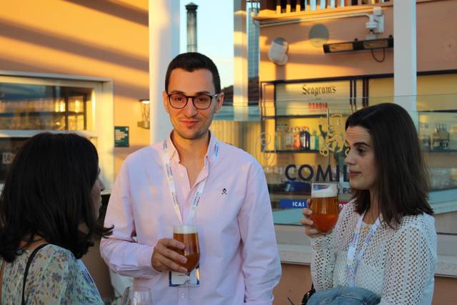 Three people having a conversation while holding drinks at an outdoor event during sunset.