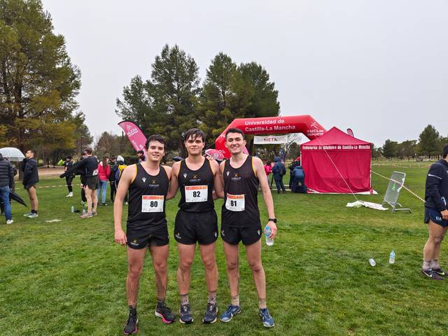 Three athletes posing together at a cross-country event, with event tents and other participants in the background.