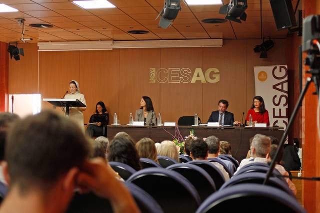Una ponente habla en un evento formal mientras una audiencia escucha atentamente.