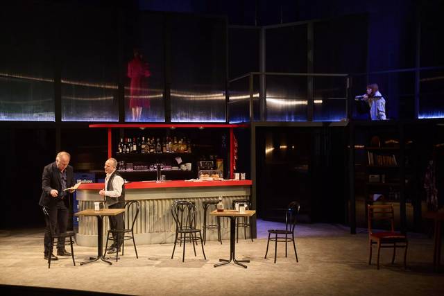Una escena de teatro que muestra un bar con dos personajes en primer plano y otros dos en balcones superiores.