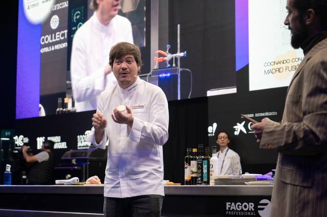 Un chef explica técnicas culinarias en un escenario ante una audiencia, con equipamiento de cocina profesional al fondo.