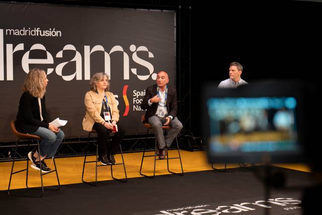 Cuatro personas sentadas participan en un panel de discusión en un evento, con una cámara desenfocada en primer plano.