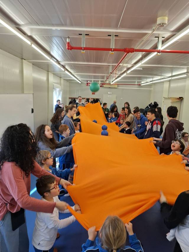 Un grupo de niños y adultos participan en una actividad recreativa usando una gran tela naranja.