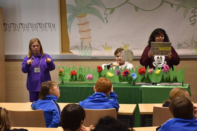 Un grupo de niños y un adulto participan en una obra de teatro con decoraciones de flores y césped artificial.