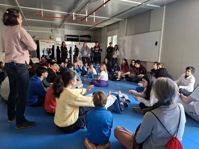 Un grupo de niños y adultos sentados en círculo, participando en una actividad en un espacio cerrado.