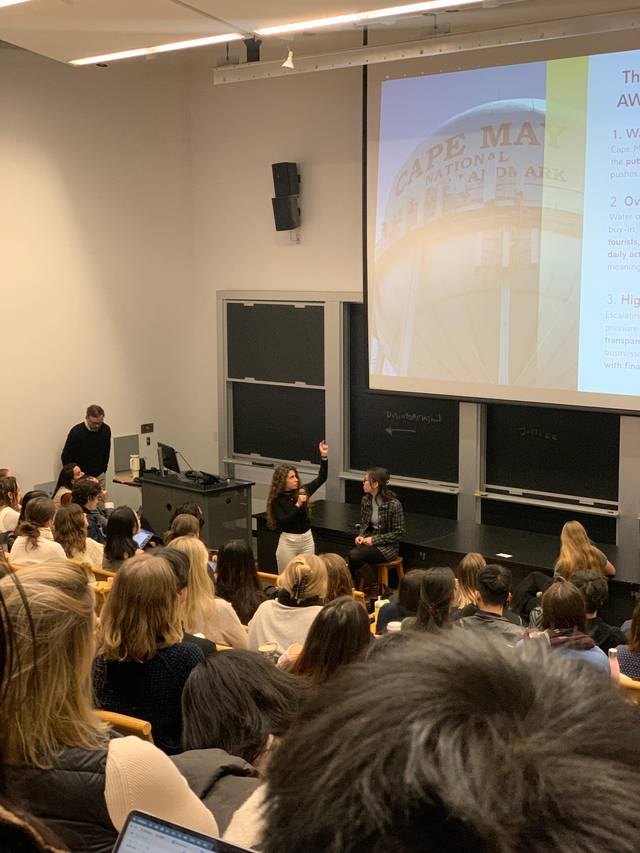 Una sala de clase llena de estudiantes atentos a una presentación frente a una pizarra.
