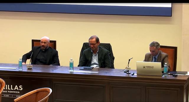 Tres hombres sentados en una mesa durante una conferencia en una sala con paneles de madera.