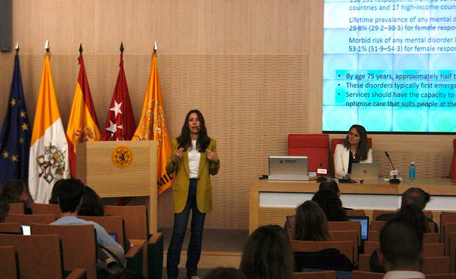 A woman delivers a presentation in a conference room with flags and audience members in the foreground.