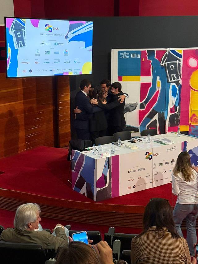 Un grupo de hombres se abraza en un evento frente a un atril y una pantalla con logotipos.
