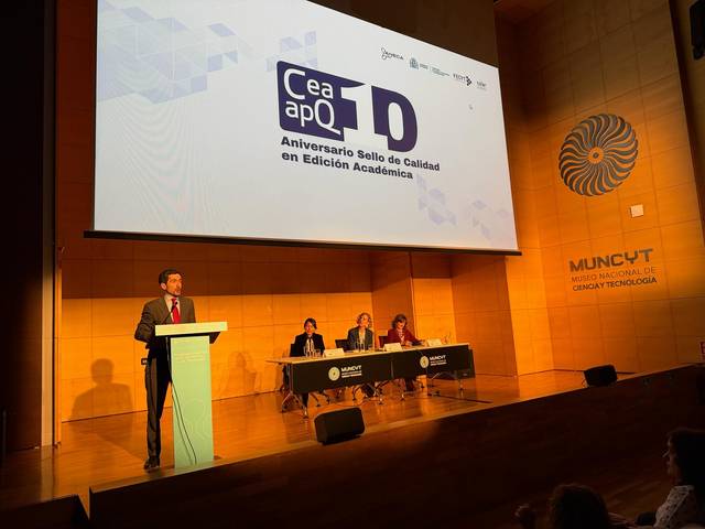Un evento con un ponente en un escenario y una mesa de panelistas en el fondo.