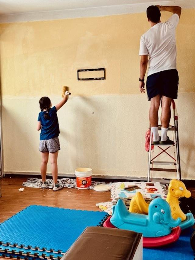 Dos personas pintando una pared en una habitación, una de pie en una escalera y otra en el suelo.