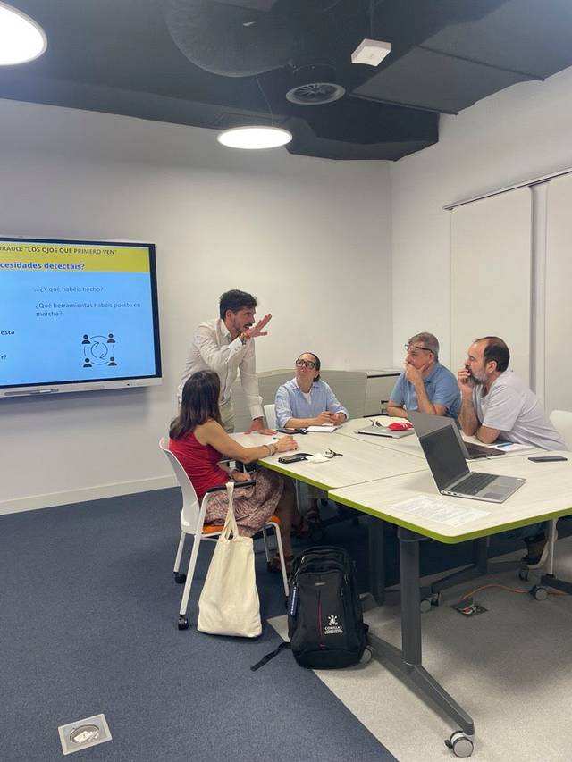 Un grupo de personas discutiendo en una sala de reuniones mientras un presentador explica una diapositiva en una pantalla.