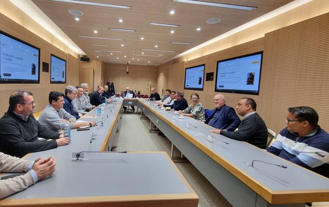 A group of people sitting around a conference table in a modern meeting room with presentations on screens.