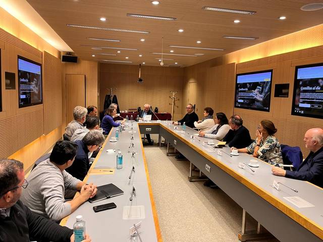 A group of people sitting at a long conference table in a modern meeting room with screens displaying a street view.