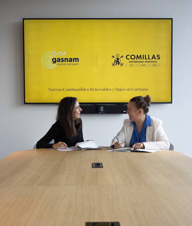 Two women are having a meeting in a conference room with a digital display in the background showing logos and text about renewable transport fuels.