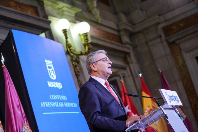 Un hombre habla en un evento con una pantalla que muestra el logo de Madrid y varios estandartes de fondo.