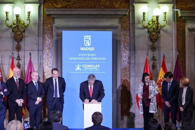 La imagen muestra un evento formal en el que varias personas están presentes alrededor de una mesa mientras un hombre firma un documento.