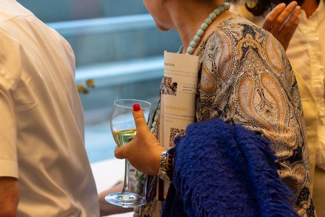A woman in a patterned dress holding a glass of white wine and a brochure at a social event.