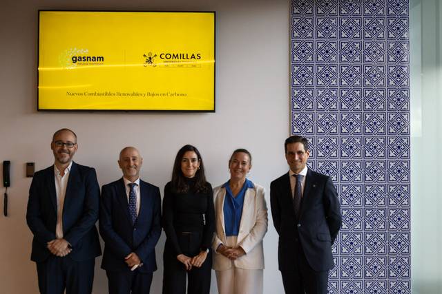 Five people standing in front of a display with the logos of Gasnam and Comillas, featuring a decorative blue tile wall on the right.