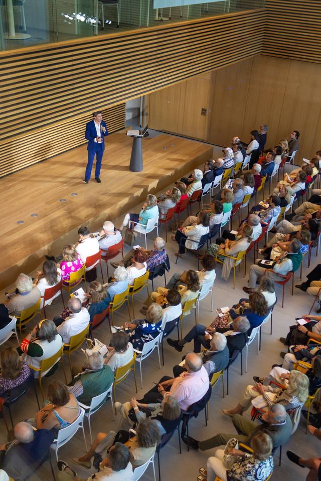 A man in a blue suit is giving a presentation to an audience seated in colorful chairs in a modern hall.
