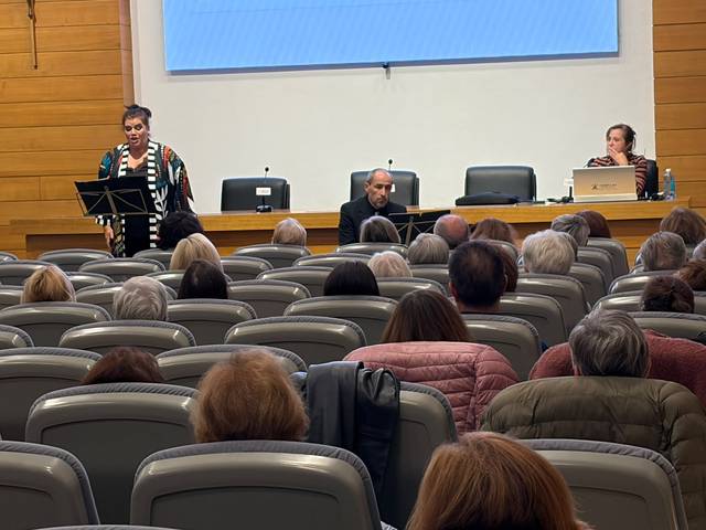 Una mujer está cantando frente a un público atento en un evento cultural.