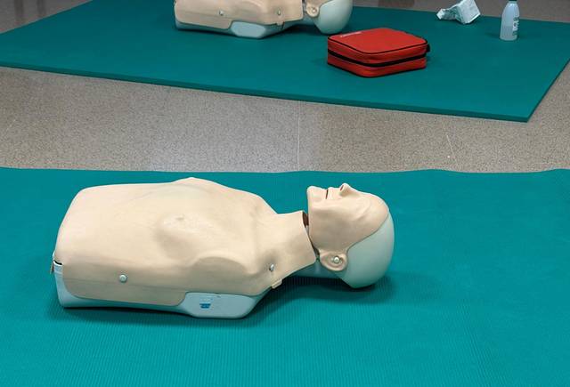 La imagen muestra un maniquí de resucitación sobre una superficie de entrenamiento verde, con un maletín rojo y otros objetos en el fondo.