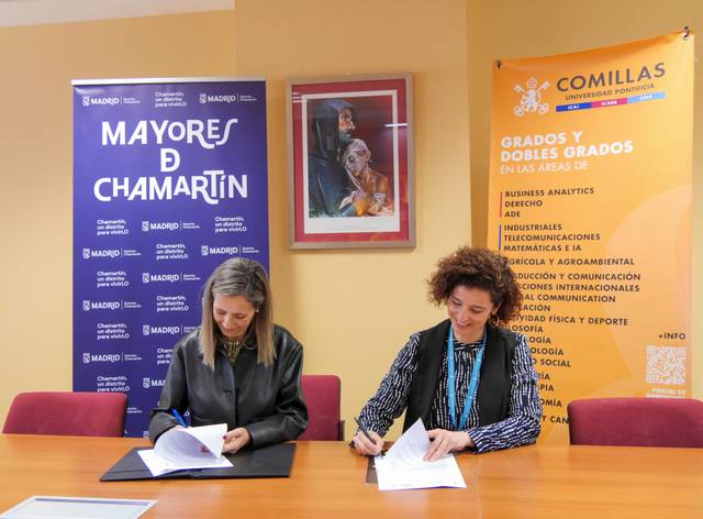 Dos mujeres están firmando documentos en un ambiente formal, rodeadas de cartelera informativa y un retrato en la pared.