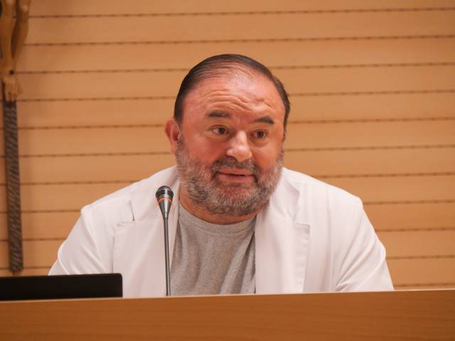 Un hombre con barba y cabello corto, vestido con una bata blanca, está hablando en un evento o conferencia.