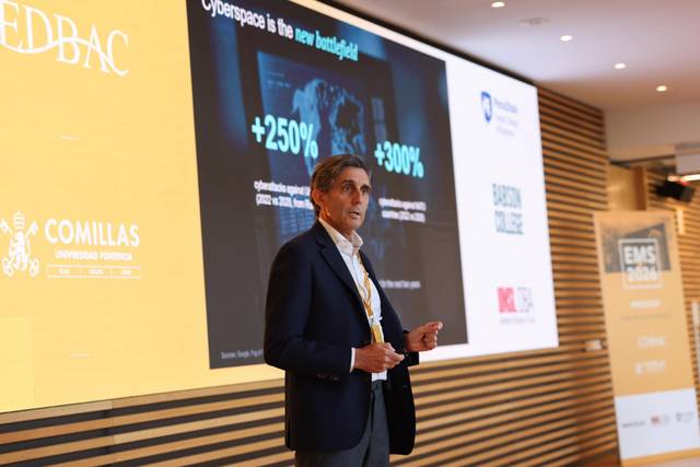 Un hombre está presentando en una conferencia, con una pantalla detrás que muestra estadísticas sobre ciberespacio.