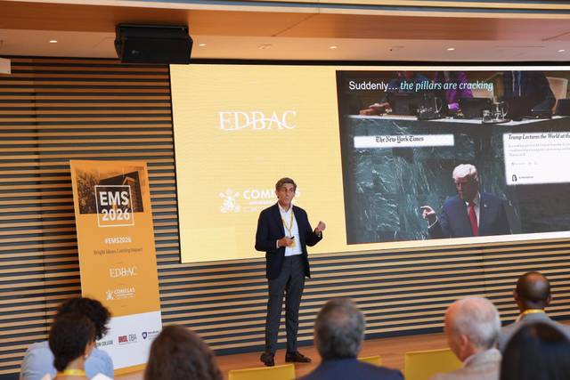 Un ponente habla sobre un tema en una conferencia mientras se proyecta información relevante en una pantalla detrás de él.