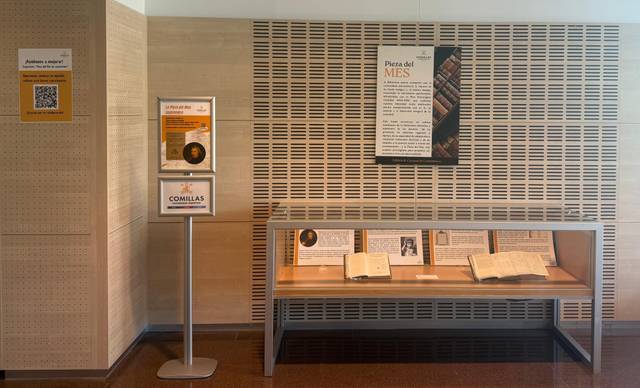 La imagen muestra un espacio dentro de un edificio con un expositor de libros y un cartel que destaca la 'Pieza del Mes'.