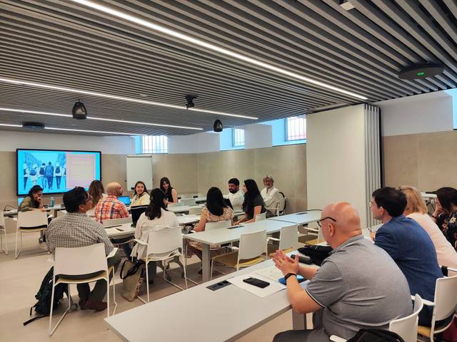 Una sala de reuniones con varias personas sentadas alrededor de mesas blancas durante una presentación.