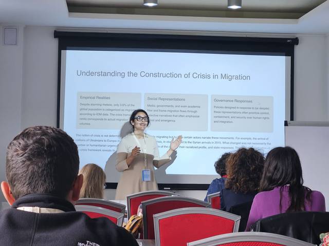 Una mujer presenta información sobre la crisis en la migración frente a una audiencia en una sala de conferencias.