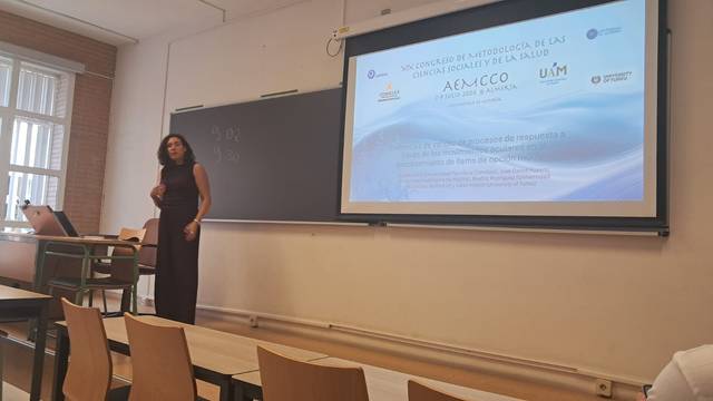 Una mujer presenta una conferencia en un aula con una proyección de diapositivas sobre metodología en ciencias de la salud.