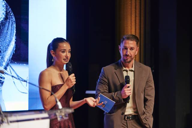 Dos presentadores están en un escenario, sosteniendo micrófonos y hablando frente a un fondo iluminado.